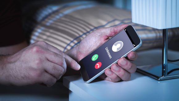 Person holding mobile and smartphone in bedroom bed home late. Phone screen reads unknown caller. 