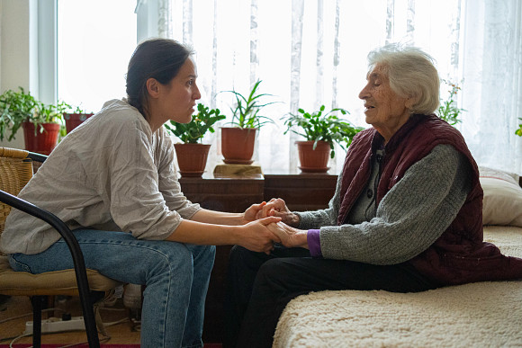 Older woman sitting on bed holding hands with a younger woman sitting in a chair. 